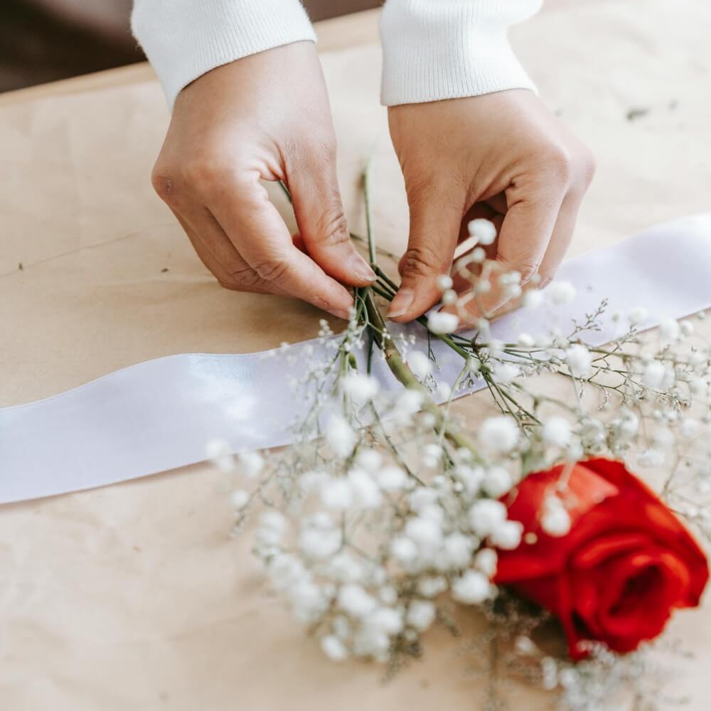Hands assembling a boutonniere using a red rose and white baby’s breath for a wedding.