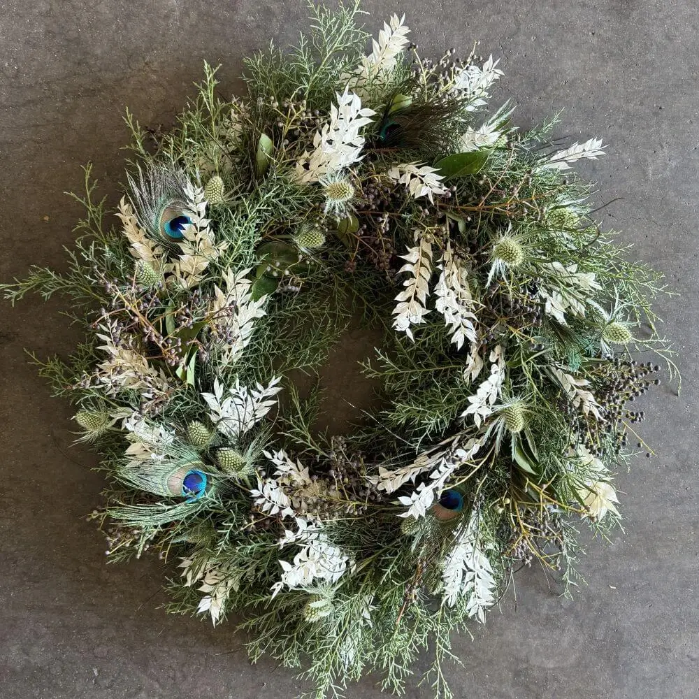 Completed DIY holiday wreath from Kit #1 featuring peacock feathers, silver eryngium, and cool-toned botanicals on grapevine base hanging display