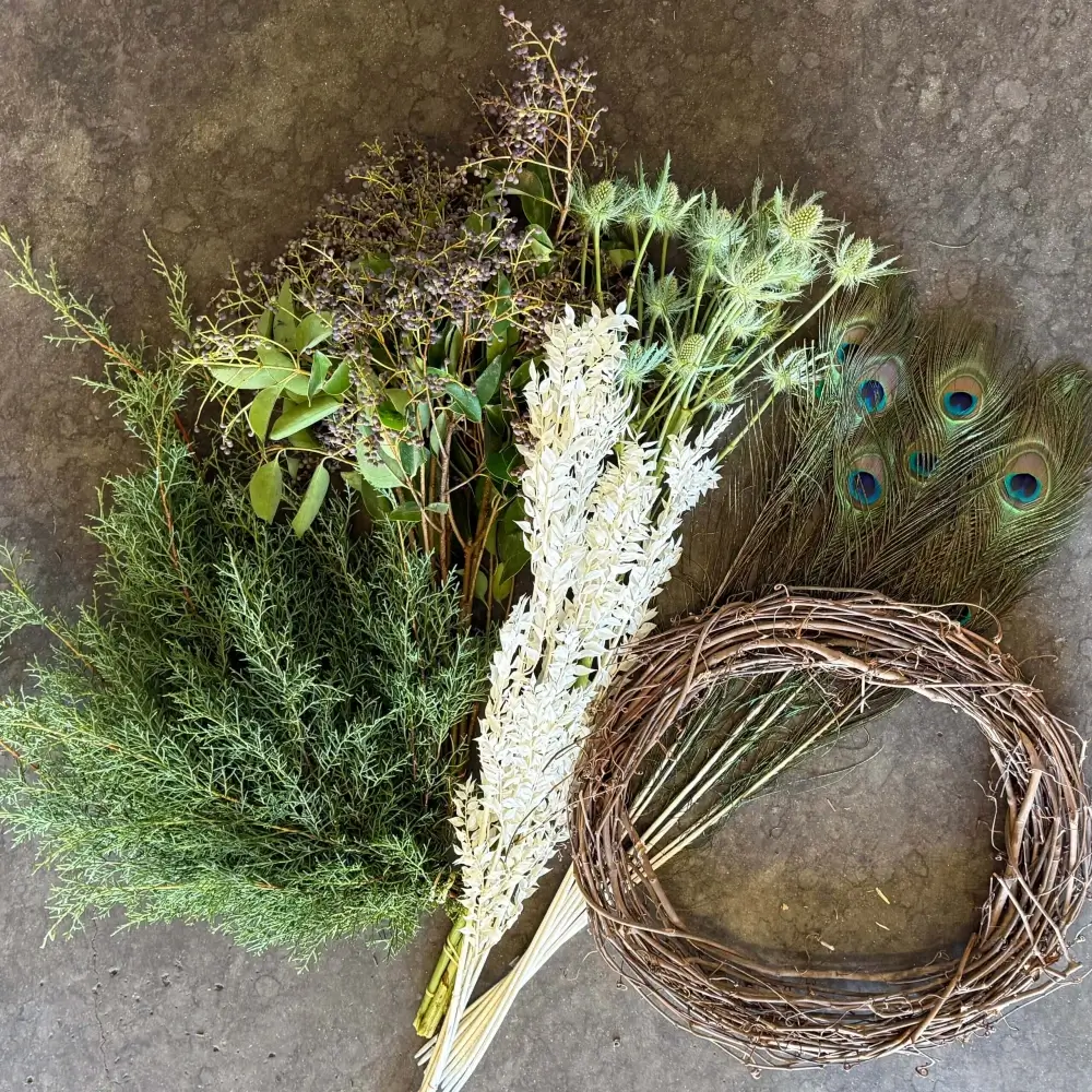 Holiday Wreath Kit #1 unassembled materials including grapevine wreath base, Carolina Sapphire, privet berry, silver eryngium, bleached Italian ruscus, and peacock feathers laid out on gray surface
