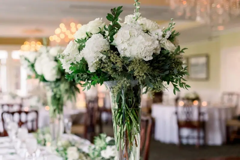 Tall glass vase centerpiece with white hydrangea, white roses, and lush green foliage arranged for a St. Patrick's Day table display