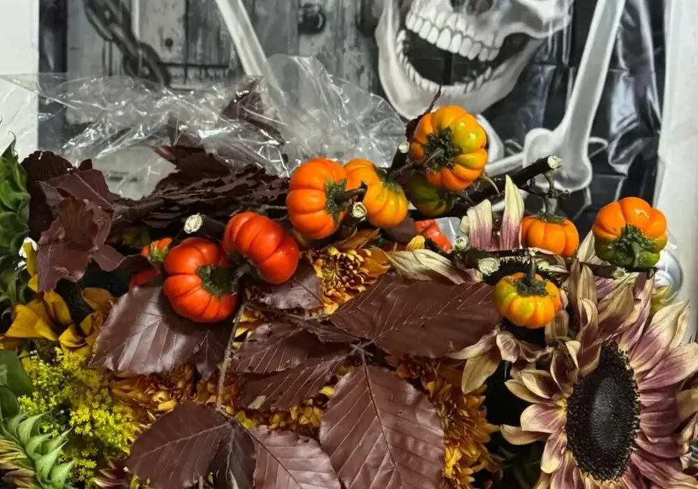 Halloween floral arrangement with orange pumpkins, burgundy sunflowers, chocolate brown foliage, and dried corn husks against skeleton backdrop